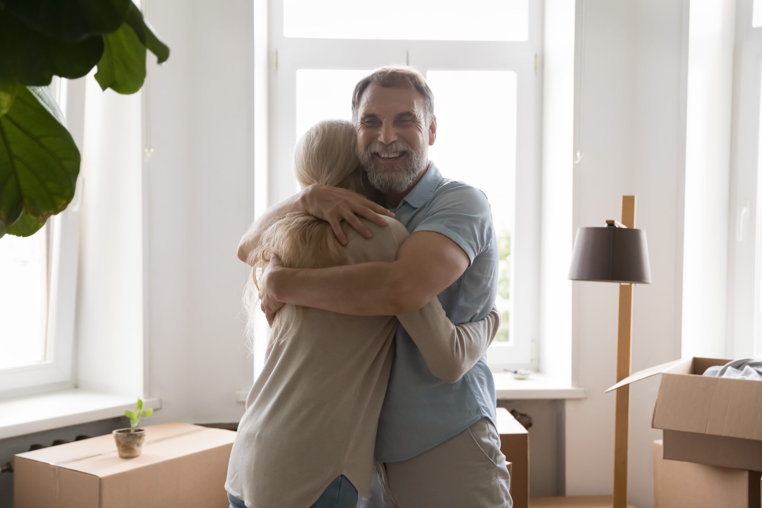 Coppia felice dai capelli grigi che si abbraccia tra scatoloni da trasloco in casa, in piedi nella nuova casa, per festeggiare un trasferimento, l'acquisto di una proprietà, la proprietà. Marito allegro che abbraccia la moglie.